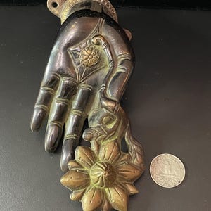 May include: A brass door knocker in the shape of a hand holding a flower. The hand is detailed with lines and the flower has a raised center.