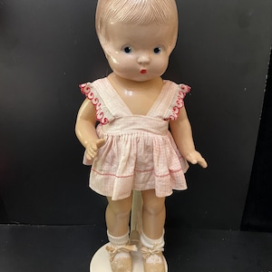 May include: A vintage porcelain doll with blonde hair, blue eyes, and a pink and white gingham dress. The doll is wearing white socks and brown shoes and is standing on a white stand.