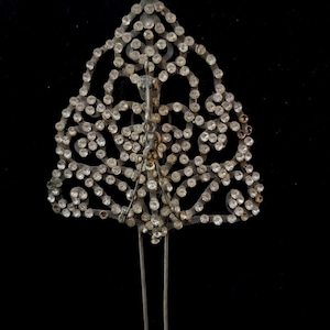 May include: A silver-toned hair pin with a decorative, openwork design in a triangular shape. The pin is embellished with numerous small, clear rhinestones. The pin has two long, parallel prongs for insertion into the hair. The background is black.