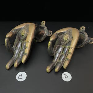 May include: Two ornate brass hand-shaped wall hooks with intricate details. The hands are in a gesture of giving or receiving, with the palms facing outward. The hooks are a dark bronze color with gold accents.