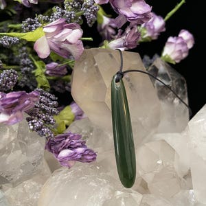 May include: A dark green jade pendant with a smooth, polished finish hangs from a black cord. The pendant is shaped like a teardrop and is resting on a bed of white quartz crystals.