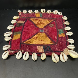 May include: A square, red, yellow, and green woven fabric with a white shell border. The fabric has a geometric pattern and a small, round, blue button in the center.