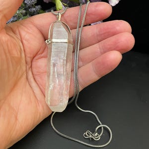 May include: A clear quartz crystal pendant on a silver chain. The crystal is long and slender, with a pointed tip. The chain is a delicate, snake-like style.