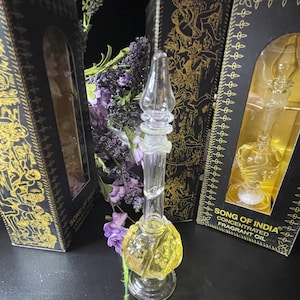 May include: A clear glass bottle with a gold liquid inside. The bottle has a decorative design and is sitting on a black surface. The bottle is labeled "Song of India Concentrated Fragrant Oil".