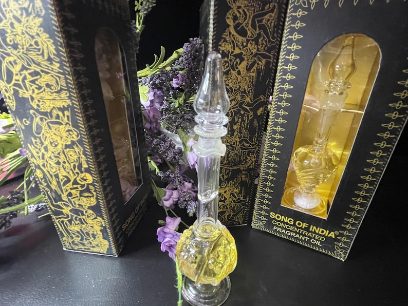 May include: A clear glass bottle with a gold liquid inside. The bottle has a decorative design and is sitting on a black surface. The bottle is labeled "Song of India Concentrated Fragrant Oil".