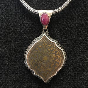 May include: A silver chain necklace with a pendant featuring a detailed floral design in gold on a dark brown background. The pendant has a small, round, pink gemstone at the top.