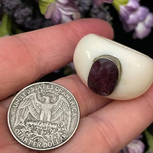 May include: A white ring with a large, faceted, dark pink gemstone set in silver. The ring is shown next to a US quarter for size comparison.