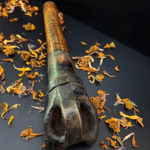 May include: A vintage, dark brown metal tool with a textured handle and a distinctive, open-ended head. The tool is set against a dark background scattered with dried orange flower petals, creating a rustic aesthetic.