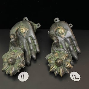 May include: Two ornate bronze hand-shaped door handles with a flower design. The handles are green and gold in color. The handles are numbered 11 and 12.