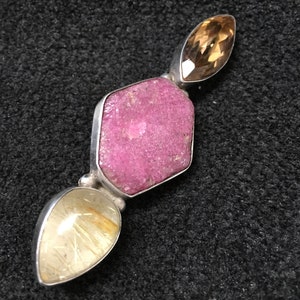 May include: A silver pendant featuring a large, irregular pink gemstone at the center, flanked by two teardrop-shaped gemstones, one golden-brown and the other a pale yellow with golden inclusions. The pendant is set against a dark, textured background.