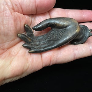 May include: A bronze hand sculpture with a detailed palm and fingers. The hand is in a giving gesture with the palm facing up. The back of the hand has a square design with a floral pattern.