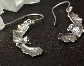 May include: A pair of silver earrings featuring a detailed bat design. The bats are facing forward with their wings spread out.