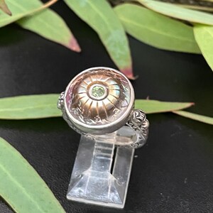 May include: A silver ring with a round, ornate setting featuring a green gemstone. The ring is on a clear display stand.
