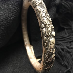 May include: Silver-toned bangle bracelet with intricate embossed floral and foliate designs. The bracelet has a hinged clasp and is set against a dark background. The detailed carvings add a vintage or antique aesthetic to the jewelry.