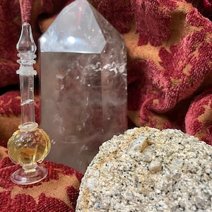 May include: A clear glass bottle with a stopper and a golden liquid inside, a large clear quartz crystal, and a rough gray rock with a white crystal embedded in it. The objects are on a red and gold patterned fabric.
