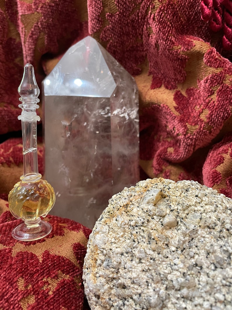 May include: A clear glass bottle with a stopper and a golden liquid inside, a large clear quartz crystal, and a rough gray rock with a white crystal embedded in it. The objects are on a red and gold patterned fabric.