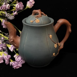 May include: A teal-colored teapot with a brown handle, spout, and lid detail. The teapot features a floral design and is set against a black background with purple flowers. The teapot's design is reminiscent of nature.