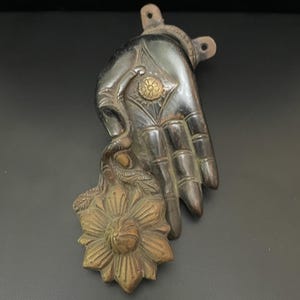 May include: A dark brown metal hand with a flower design, holding a gold metal flower with a raised center. The hand has a diamond-shaped design on the palm and three holes on the back.