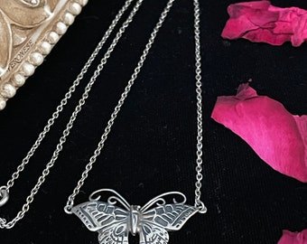 May include: A silver butterfly necklace with a delicate chain. The butterfly pendant is detailed and intricate, with a textured surface. The necklace is photographed on a black background with pink rose petals.