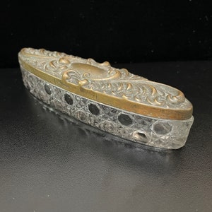 May include: A vintage, oval-shaped trinket box with a clear glass bottom and a brass top with an ornate floral design.
