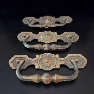 May include: Three antique brass drawer pulls with a floral design in the center. The pulls have a curved handle and are set against a black background. The brass has a slightly aged, tarnished appearance.