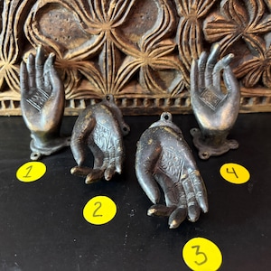 May include: Four antique bronze-toned hand sculptures. Two are upright with bases, and two are pendant-style. The hands are detailed with etched patterns and have a weathered appearance. The background features carved wooden panels.