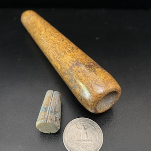 May include: A brown and tan wooden pipe with a carved design. The pipe is about 6 inches long and has a diameter of about 1 inch. The pipe is next to a small piece of wood and a US quarter for scale.