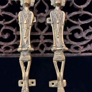 May include: A pair of matching brass door handles, each shaped like a stylized human figure with detailed facial features, necklaces, and textured waistbands. The figures have elongated legs and arms, with mounting plates at the top and bottom for installation.
