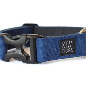 Puede incluir: Un collar azul marino para perros con hebilla de plástico negro y anilla en D de metal plateado. El collar tiene una etiqueta negra con el texto "KIWI DOGS" en blanco. El collar está hecho de tela texturizada.