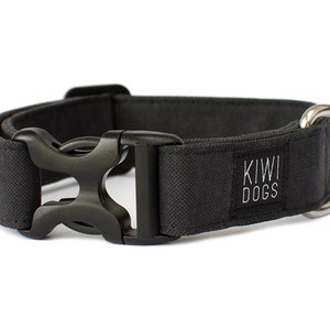 Puede incluir: Un collar negro para perros con una hebilla de plástico negro y una anilla en D de metal plateado. El collar tiene el texto "KIWI DOGS" bordado en blanco sobre una etiqueta de tela negra.