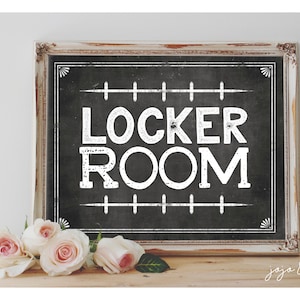 Puede incluir: Un cartel blanco con letras negras que dice "Locker Room" en una fuente vintage. El cartel está enmarcado en un marco blanco desgastado y está sentado sobre una superficie de madera con rosas rosadas delante.