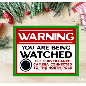 May include: A red and green warning sign with white text that reads "WARNING YOU ARE BEING WATCHED ELF SURVEILLANCE CAMERA CONNECTED TO THE NORTH POLE THIS AREA UNDER 24 HOUR LIVE VIDEO RECORDED SURVEILLANCE". The sign has a black and white image of a camera lens.