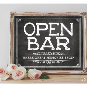 Puede incluir: Un letrero de pizarra enmarcado en blanco con el texto "OPEN BAR" en letras blancas. El letrero también incluye el texto "WHERE GREAT MEMORIES BEGIN" en letras blancas. El letrero está sentado sobre una superficie de madera con rosas rosadas delante.