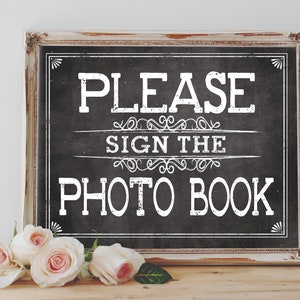 May include: A white sign with a black chalkboard background in a distressed white frame. The sign says "Please sign the photo book" in white text with decorative flourishes.