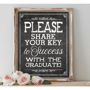 May include: A white framed chalkboard sign with black text that reads "Please share your key to success with the graduate!" The sign is sitting on a wooden surface with pink roses in front of it.