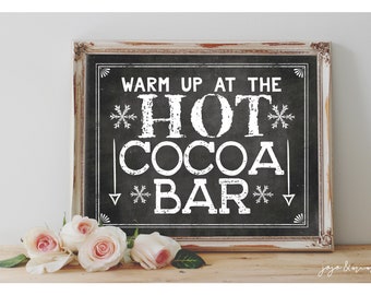May include: A white sign with black lettering in a rustic wooden frame. The sign reads "Warm up at the Hot Cocoa Bar" with snowflakes and arrows around the text. The sign is sitting on a wooden surface with pink roses in front of it.
