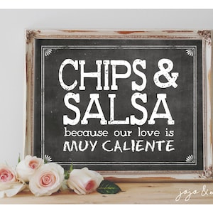 May include: A framed chalkboard sign with white text that reads "Chips & Salsa because our love is muy caliente". The sign is sitting on a wooden surface with pink roses in front of it.