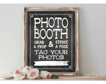 May include: A framed chalkboard sign with the words "PHOTO BOOTH" in large white letters. Below, it reads "GRAB A PROP & STRIKE A POSE TAG YOUR PHOTOS!". The frame is distressed white, and roses are at the bottom.