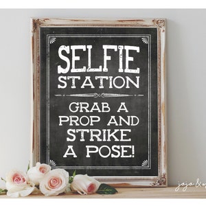 May include: A white framed chalkboard sign with black lettering that reads "Selfie Station Grab a Prop and Strike a Pose!" The sign is sitting on a wooden surface with pink roses in front of it.