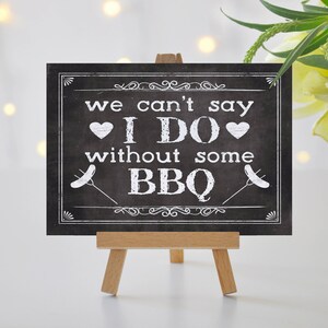 May include: Black chalkboard sign with white text that reads "We can't say I DO without some BBQ". The sign is on a small wooden easel.