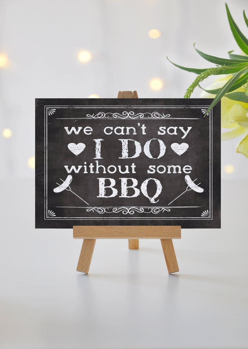 May include: Black chalkboard sign with white text that reads "We can't say I DO without some BBQ". The sign is on a small wooden easel.