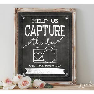Puede incluir: Un letrero de pizarra enmarcado en blanco con texto negro que dice "Help us capture the day" con un gráfico de cámara estilizado y el texto "Use the hashtag" debajo. El letrero está sobre una superficie de madera con algunas rosas rosadas delante.