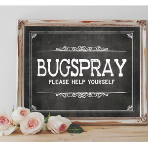 Puede incluir: Un letrero de pizarra enmarcado en blanco con el texto "BUGSPRAY PLEASE HELP YOURSELF" en letras blancas. El letrero está sobre una superficie de madera con rosas rosadas delante.