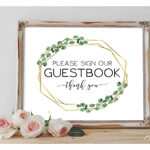 May include: A white sign with a gold geometric frame and green eucalyptus leaves around the frame. The sign says "Please sign our guestbook - thank you".