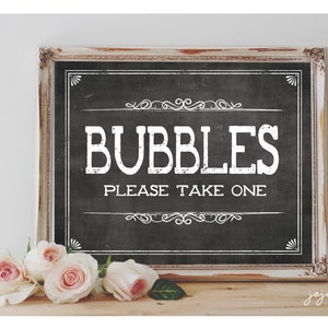 May include: A white sign with black lettering in a distressed wood frame. The sign reads "Bubbles Please Take One" with a decorative border. The sign is sitting on a wooden surface with pink roses in front of it.
