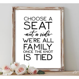 Op de afbeelding: Een wit ingelijst bord met zwarte tekst die luidt: "Kies een stoel, niet een kant. We zijn allemaal familie zodra de knoop gelegd is." Het bord staat op een houten oppervlak met roze rozen ervoor.