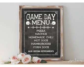 May include: A white framed chalkboard sign with the text "GAME DAY MENU" in black lettering. The menu includes pizza, nachos, homemade chili, hot dogs, hamburgers, and corn dogs. The sign also states "ALSO SERVING REFRESHMENTS".