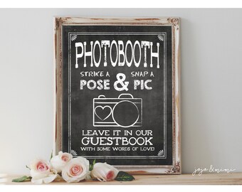May include: A white framed chalkboard sign with the text "PHOTOBOOTH STRIKE A POSE & SNAP A PIC LEAVE IT IN OUR GUESTBOOK WITH SOME WORDS OF LOVE" and a black and white illustration of a camera with a heart.
