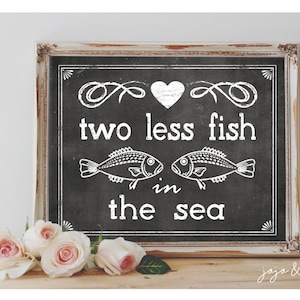 Pode incluir: Uma lousa preta com moldura branca, com o texto "two less fish in the sea" com um coração e duas ilustrações de peixes. A lousa está sobre uma superfície de madeira com rosas cor-de-rosa na frente.