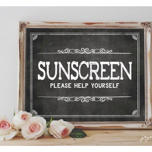 Puede incluir: Un letrero de pizarra enmarcado con texto blanco que dice "SUNSCREEN PLEASE HELP YOURSELF". El letrero está sobre una superficie de madera con rosas blancas delante.